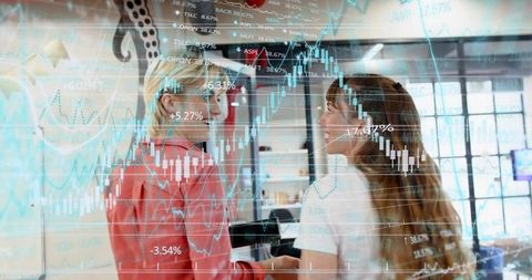 Two professional women analyzing candlestick charts on glass, collaborating in modern office