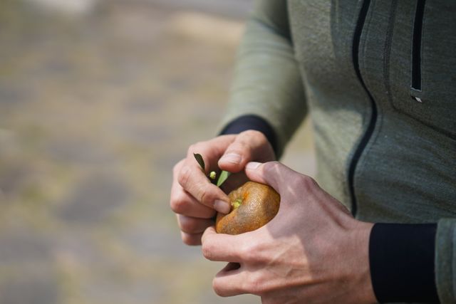 Person wearing a green hoodie is peeling a fresh orange outside. This can be used for concepts related to healthy eating, outdoor activities, casual attire, and preparing fruit. Ideal for nutritional blogs, lifestyle content, and wellness promotion.