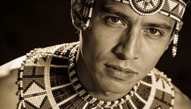 Gazing male wearing beaded geometric headdress and broad collar sepia portrait