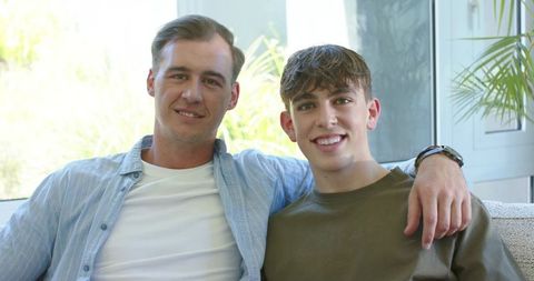 Father and teen son sitting on sunlit couch, arm draped around shoulder, smiling together