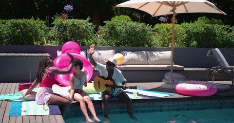 African american friends lounging poolside high-fiving with guitar and donut float