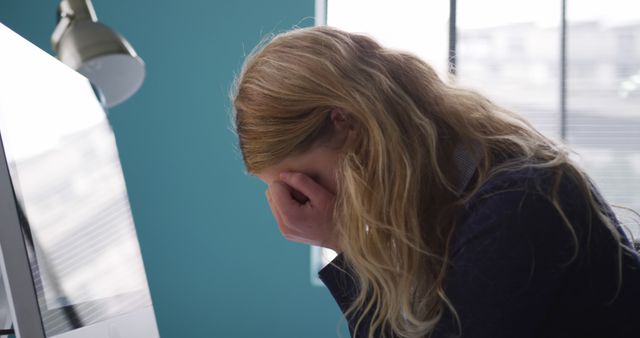 Young woman expressing stress and fatigue in office setting, head resting on hand near computer monitor. Useful for articles on workplace stress, corporate burnout, mental health, and work-life balance.