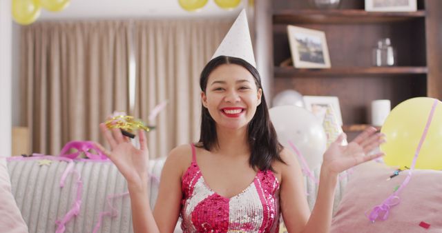 Young woman enjoys birthday party at home with festive decorations, balloons, and confetti. She is smiling and dressed in a colorful outfit with a party hat. Great for themes related to celebrations, birthdays, happiness, home parties, and joyful moments.