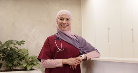Smiling female nurse wearing hijab and burgundy scrubs leaning on clinic reception counter