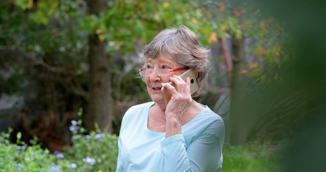 Senior Caucasian woman talks on her phone in a garden. She seems engaged in a conversation amidst the tranquility of an outdoor setting.