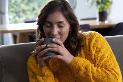Young woman in her 20s inhaling warm drink aroma in mustard knit sweater on sofa