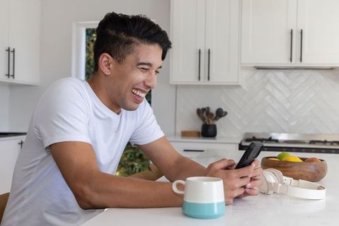 African american man smiling and using smartphone at modern kitchen island with mug