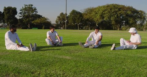 Diverse cricket teammates sitting on grass relaxing in white kit during sunny afternoon