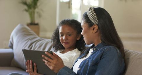 Diverse mother and daughter sharing tablet on sofa, casual denim and cozy living room moment