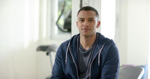 Young man sitting in home gym wearing navy hoodie and grey tee near exercise bike, bright studio