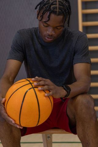 African american basketball player sitting bench holding orange ball wearing red shorts