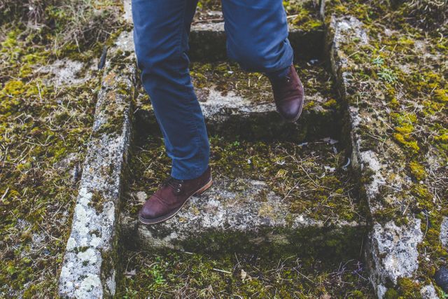 Person wearing brown boots and blue pants stepping down an old stone staircase covered with moss in outdoor setting. Image depicting themes of exploration, adventure, and nature's reclaiming of man-made structures. Useful for travel websites, outdoor activity promotions, and nature conservation campaigns.