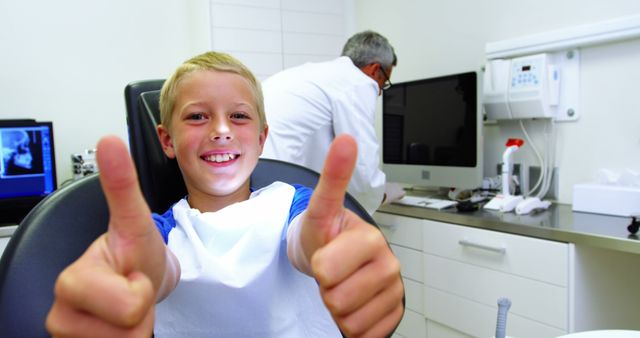 A young boy with light hair, smiling widely, gives two thumbs up while sitting in a dental chair at a modern dental clinic. The dentist, in a white coat, is seen in the background preparing equipment. This image could be used for promoting pediatric dental services, dental health awareness in children, or advertisements for dental clinics.