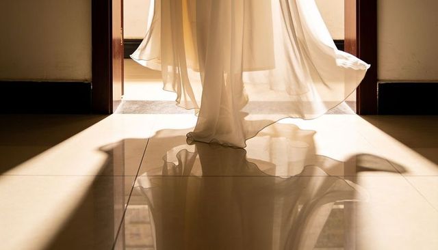 Woman wearing sheer gown flowing through sunlit doorway with mirror-like reflection