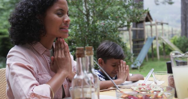 Mother and son are praying before sharing a meal at an outdoor table in a lush garden. The setting suggests a peaceful family moment filled with love and faith. This can be used to depict family bonding, gratitude during meals, faith practices, or peaceful summer moments. Suitable for themes centered around family values, spirituality, and outdoor lifestyle.
