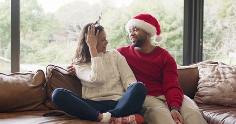 Couple sitting cozily on couch wearing santa hat and reindeer headband, festive home moment