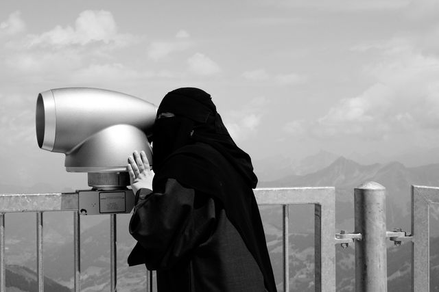 A woman in traditional attire using a telescope on a viewing platform with a mountainous background. The image is in black and white, highlighting cultural aspects and offering a picturesque view of the mountains. Ideal for themes related to travel, sightseeing, adventure, and cultural exploration. It can be used for travel blogs, tourism websites, cultural preservation articles, or photography exhibits.