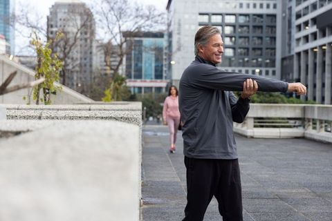 Senior man stretching on urban terrace with woman walking in background