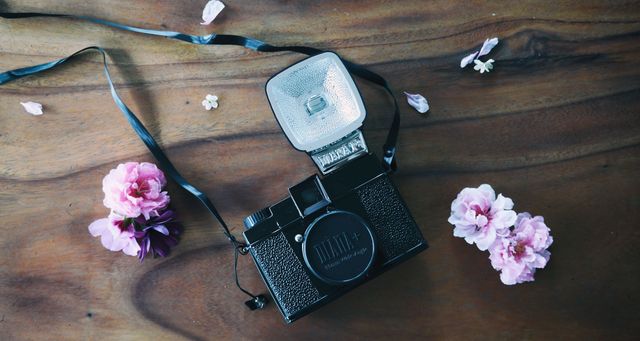Wooden table adorned with a vintage camera and pink flowers creates a nostalgic and artistic atmosphere. Perfect for use in photography-related promotions, retro-themed projects, creative blogs, or decorating a photographer’s space with a touch of elegance.