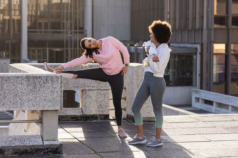Two diverse women stretching on sunlit rooftop parapet in urban athleisure