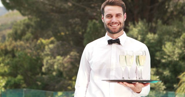 A young Caucasian male waiter is serving champagne outdoors, with copy space. His professional attire and friendly demeanor contribute to the welcoming atmosphere of the event.