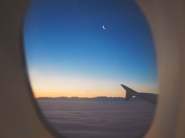 View from airplane window showing crescent moon at twilight over clouds. Useful for travel advertisements, airline promotions, inspirational articles, and websites focused on aviation or sky photography.