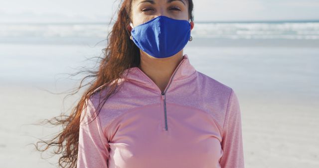 Young woman standing on a beach wearing a blue face mask. She is wearing a pink top and has the ocean and sandy shore in the background. Perfect for themes related to health, travel, recreation, safety, and outdoor activities.