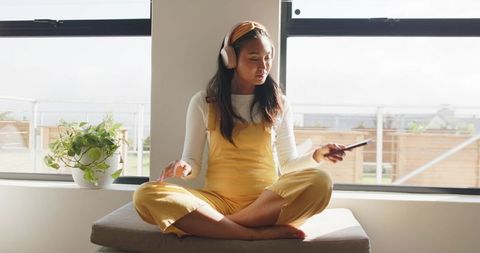 Asian woman relaxing on bench by sunny window listening to music with smartphone