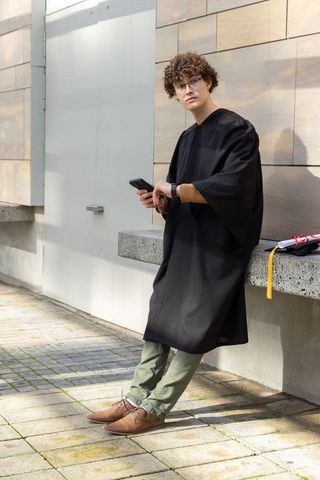Curly male graduate wearing black gown leaning on bench using smartphone with mortarboard
