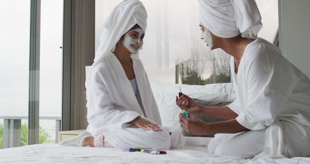 Women enjoying a spa day, wearing white bathrobes and towels on head, relaxed on bed while applying facial masks. Ideal for skincare, wellness, and beauty content. Suitable for promoting spa services, self-care products, and relaxation techniques featuring leisure and pampering sessions.