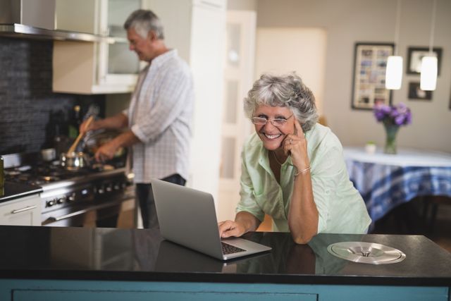 Portrait of smiling senior woman using laptop while husband cooking in kitchen