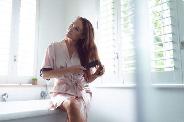 Woman combing her hair while sitting on the edge of bathtub in bathroom at home