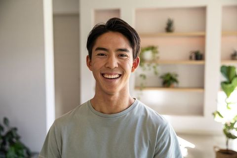 Smiling asian man standing in bright minimalist living room with built-in wooden shelves