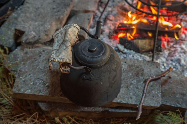 Rustic blackened teapot sitting above open campfire outdoors, boiling water narrative. Perfect for themes of wilderness adventure, traditional camping, survival skills, outdoor cooking. Use in articles and advertisements about outdoor gear, survival tips, camping recipes, and adventure travel.