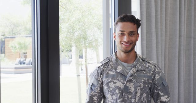 Young military officer standing indoors by window, smiling confidently in army uniform. Ideal for themes of patriotism, military service, dedication, and recruitment campaigns.