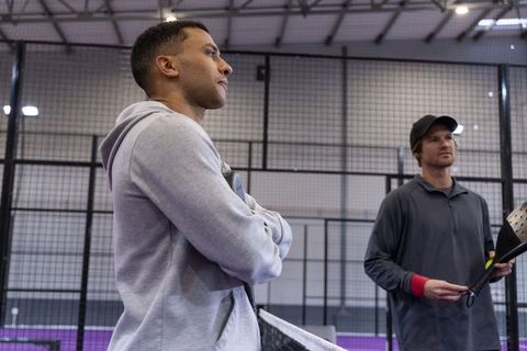 Focused padel players standing by net on indoor purple court wearing hoodie and cap