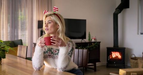 Blonde woman wearing festive sweater enjoying hot drink by fireplace during cozy christmas