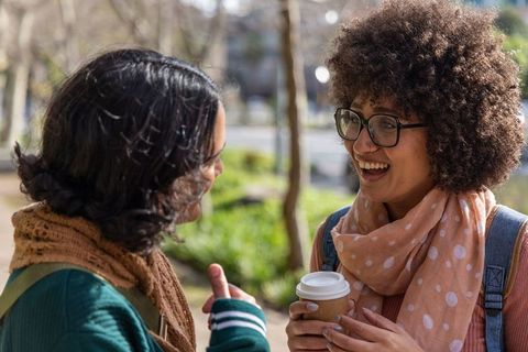 Friends chatting and laughing while holding coffee on sunny campus walkway