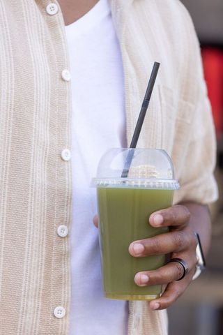 African man holding green cold-pressed juice cup with black straw in beige shirt, urban cafe
