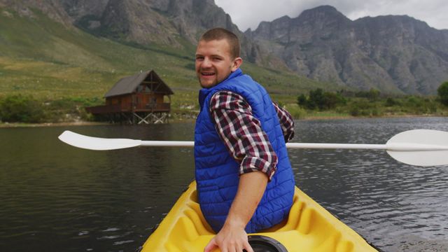 A man in a plaid shirt and blue vest enjoys a kayaking trip on a calm lake with mountains in the background. He is looking over his shoulder toward the camera, smiling, capturing a moment of leisure and joy in a picturesque setting. This image is suitable for tourism advertisements, travel blogs, or outdoor sports promotions, drawing attention to nature exploration and adventurous vacations.