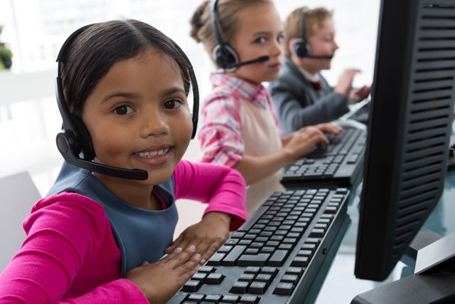 This image shows a young girl smiling while working as a customer care executive in an office setting. She is wearing a headset and using a computer keyboard. Two other children are also working in the background, indicating teamwork and a collaborative environment. This image can be used to represent concepts of young professionals, customer service, communication, and diversity in the workplace.