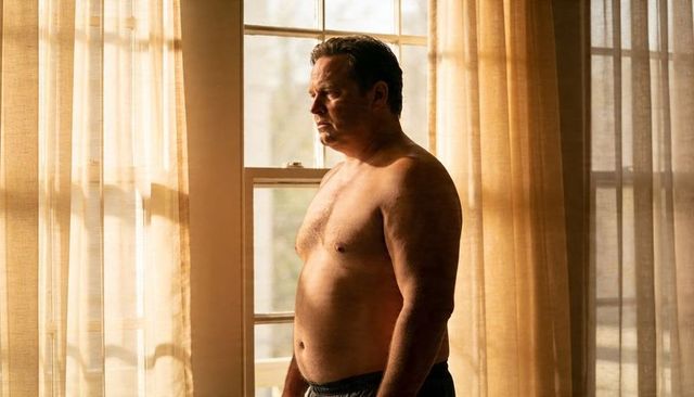 Middle-aged man standing shirtless gazing through sunlit sheer curtains at window