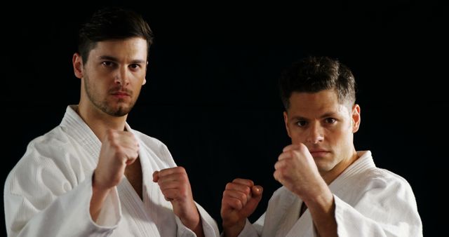 Two male martial artists are posing in fighting stances while dressed in white karate uniforms. The serious expressions on their faces show their focus and determination. This image can be used for promoting martial arts classes, illustrating articles on self-defense, and showcasing discipline and training in sports.