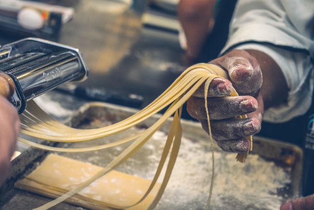Hands of a chef holding fresh pasta strands, dusted with flour, while using a pasta machine on a kitchen countertop. Great for content about cuisine, cooking tutorials, culinary arts, traditional Italian food, and homemade pasta recipes.