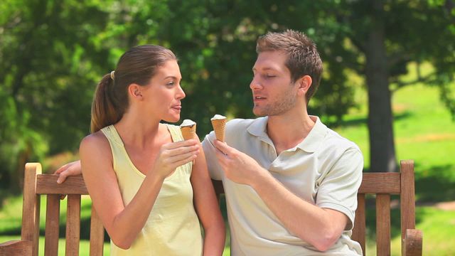 Couple sitting on comfortable wooden bench in lush green park, savoring ice creams on a sunny day. This content symbolizes leisure, carefree enjoyment, and connection in natural surroundings. Ideal for lifestyle blogs, advertisements promoting outdoor activities, summer events, or articles on relationship bonding.