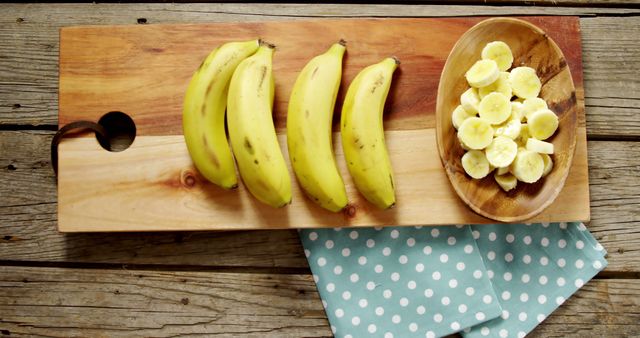 Whole bananas and sliced bananas are presented on a wooden cutting board, with copy space. A polka-dotted cloth adds a touch of homeliness to the food preparation scene.