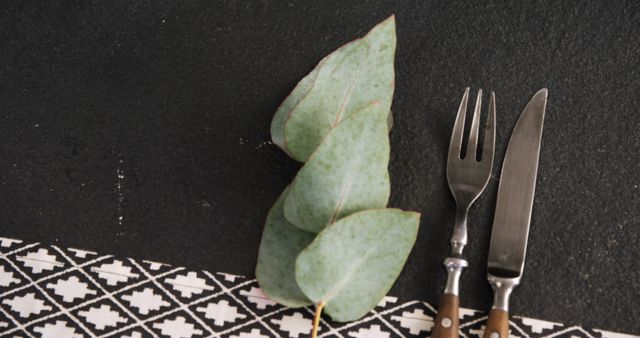 A leaf is placed next to a fork and knife on a patterned tablecloth, with copy space. The arrangement suggests a focus on natural elements in dining or an eco-friendly meal concept.