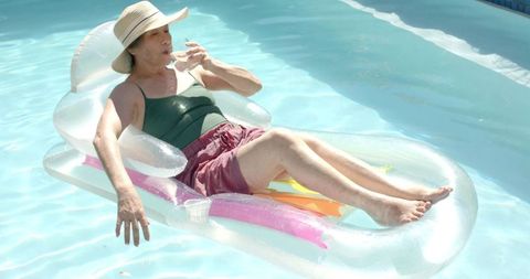 Mature asian woman relaxing on clear inflatable pool lounger sipping water under sun hat