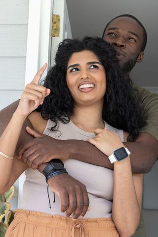 Black man embracing south asian woman in doorway, woman pointing up and showing smartwatch