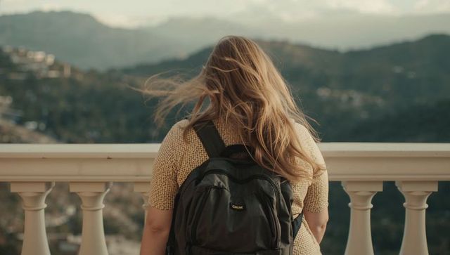 Standing woman gazing over misty mountain valley from balustrade, backpack and windblown hair